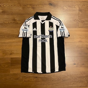 May include: A black and white striped soccer jersey with the text "northern rock" on the front. The jersey has a collar and short sleeves with black stripes. The Adidas logo is visible on the chest.