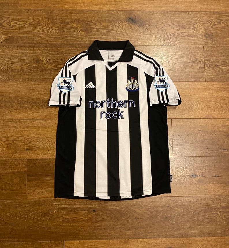 May include: A black and white striped soccer jersey with the text "northern rock" on the front. The jersey has a collar and short sleeves with black stripes. The Adidas logo is visible on the chest.