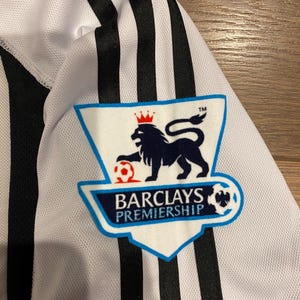 May include: Close-up of a white and black striped jersey. The jersey features the Barclays Premiership logo on the upper left chest. The logo is a blue and white shield with a lion and soccer ball.