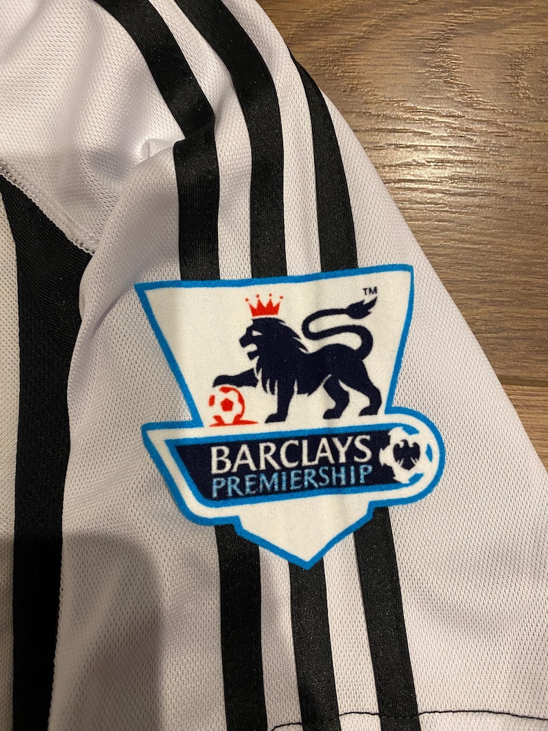 May include: Close-up of a white and black striped jersey. The jersey features the Barclays Premiership logo on the upper left chest. The logo is a blue and white shield with a lion and soccer ball.