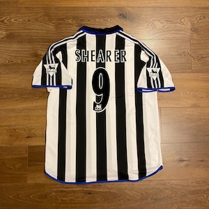May include: A black and white striped soccer jersey with the name "SHEARER" and the number "9" on the back. The jersey has blue trim and the Premier League logo on the sleeves.