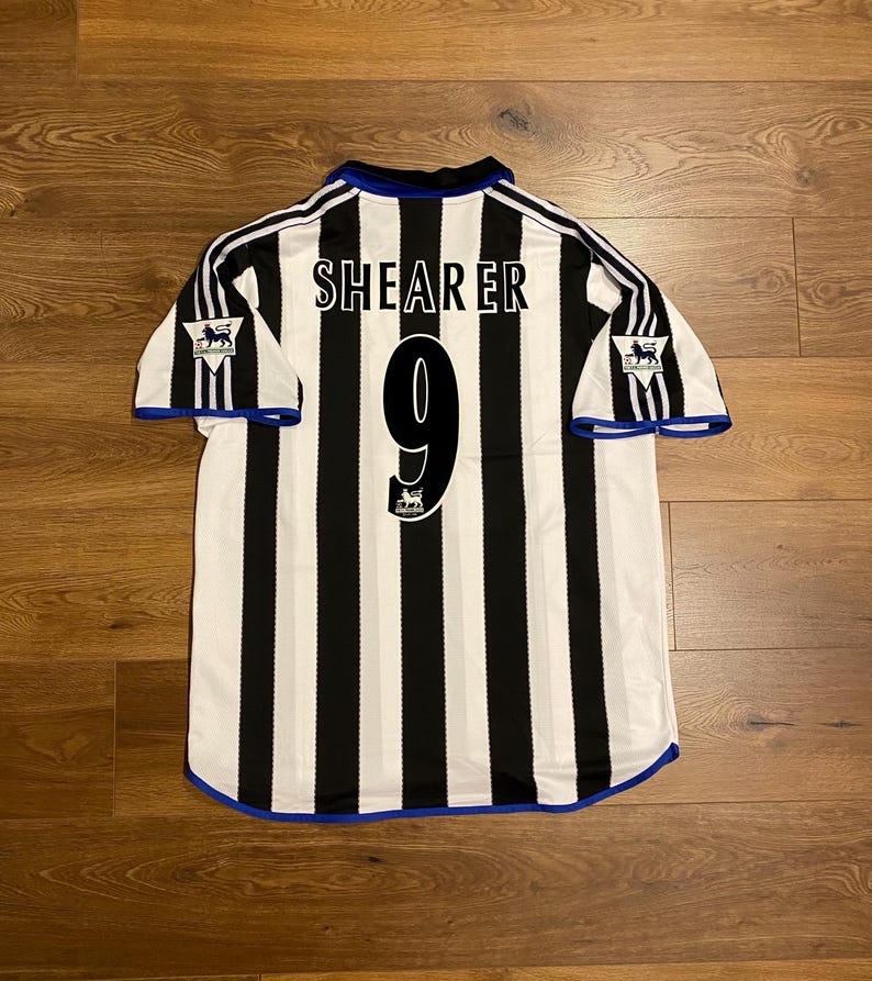 May include: A black and white striped soccer jersey with the name "SHEARER" and the number "9" on the back. The jersey has blue trim and the Premier League logo on the sleeves.