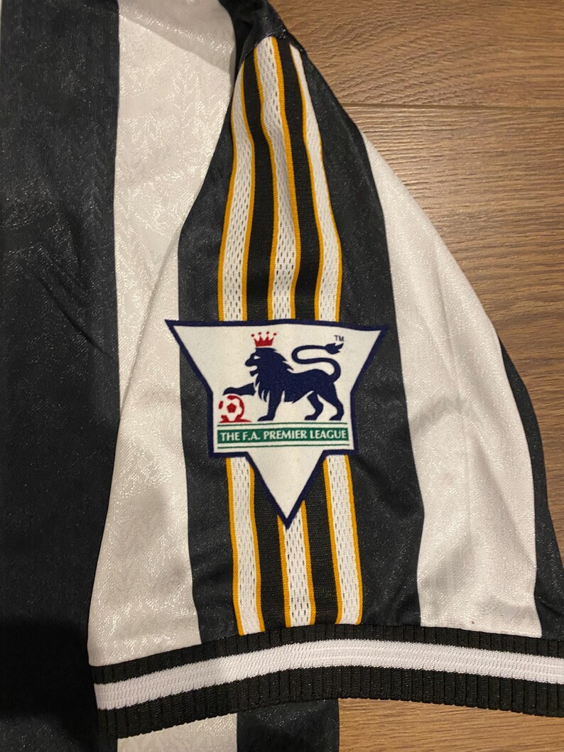 Może przedstawiać: Czarno-biała koszulka piłkarska w paski z białym rękawem i logo Premier League. Rękaw ma czarne, białe i złote paski. Koszulka jest wykonana z błyszczącego materiału.