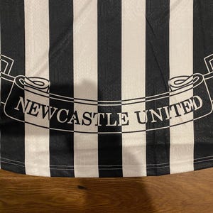 Może przedstawiać: Czarno-biała koszulka piłkarska w paski z napisem "NEWCASTLE UNITED" na banerze. Koszulka ma klasyczny design z naprzemiennymi czarno-białymi pionowymi paskami.