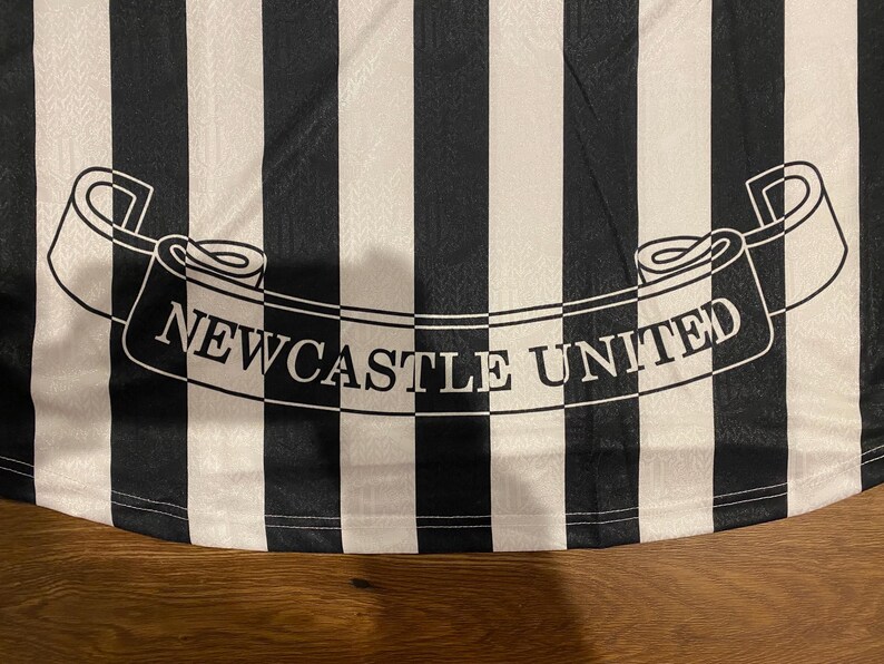 Może przedstawiać: Czarno-biała koszulka piłkarska w paski z napisem "NEWCASTLE UNITED" na banerze. Koszulka ma klasyczny design z naprzemiennymi czarno-białymi pionowymi paskami.