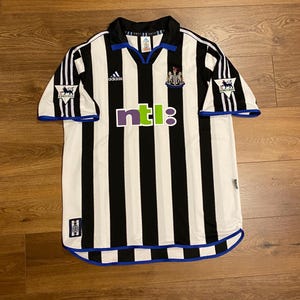 May include: A black and white striped soccer jersey with blue accents. The jersey features the Adidas logo, the Newcastle United crest, and the NTL logo. The sleeves have a Premier League logo. The collar and cuffs are trimmed in blue.