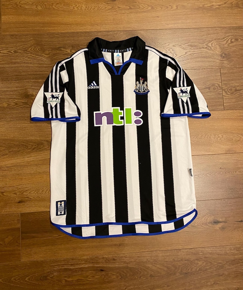 May include: A black and white striped soccer jersey with blue accents. The jersey features the Adidas logo, the Newcastle United crest, and the NTL logo. The sleeves have a Premier League logo. The collar and cuffs are trimmed in blue.