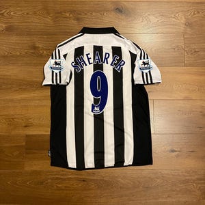 May include: Black and white striped soccer jersey with the name "SHEARER" and the number "9" in blue. The jersey has a white collar and black stripes on the sleeves. The Barclays Premier League logo is on the sleeves.