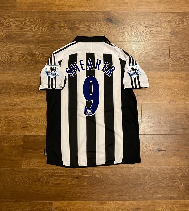 May include: Black and white striped soccer jersey with the name "SHEARER" and the number "9" in blue. The jersey has a white collar and black stripes on the sleeves. The Barclays Premier League logo is on the sleeves.