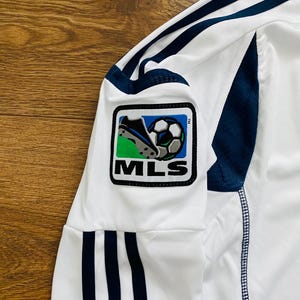 May include: White soccer jersey with navy blue stripes and accents. The jersey features an MLS logo patch on the sleeve, showcasing a soccer ball and cleats. The fabric appears to be a lightweight, athletic material.