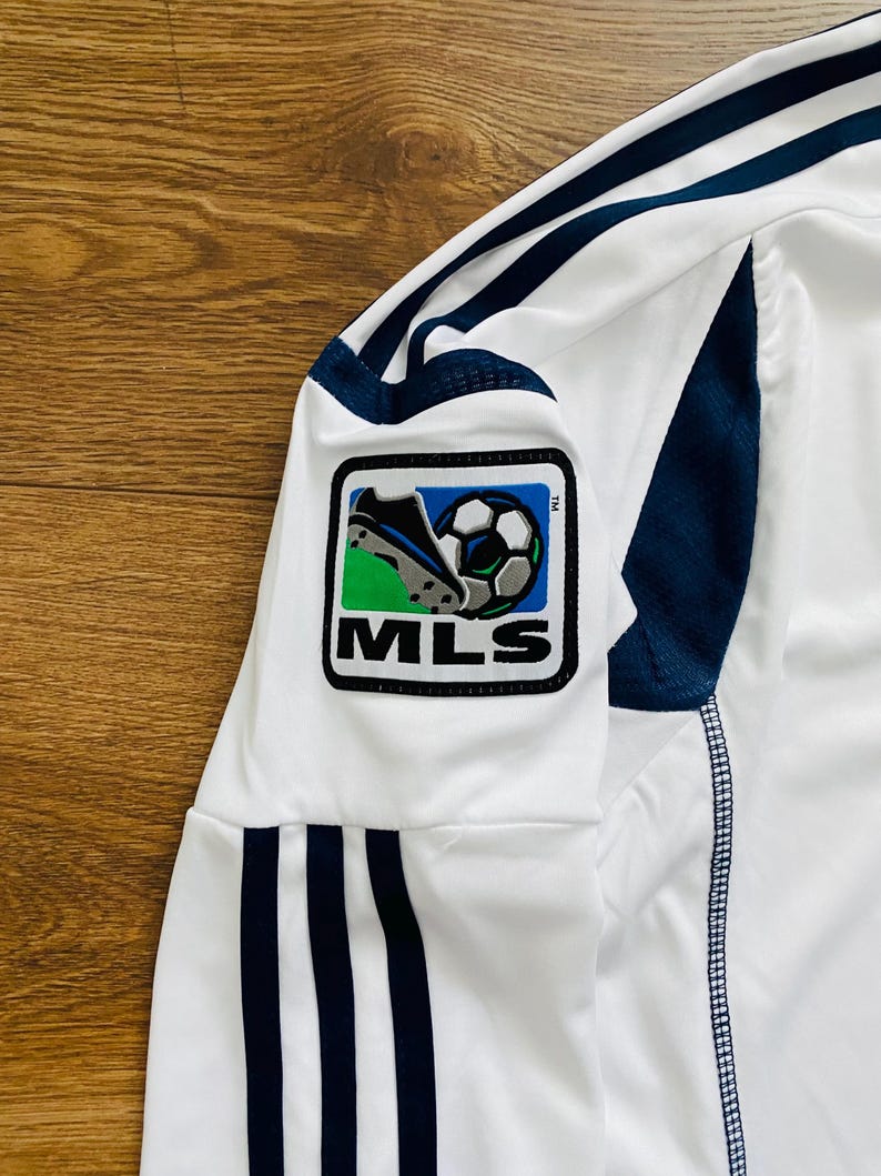 May include: White soccer jersey with navy blue stripes and accents. The jersey features an MLS logo patch on the sleeve, showcasing a soccer ball and cleats. The fabric appears to be a lightweight, athletic material.