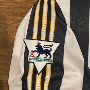Może przedstawiać: Czarno-biała koszulka piłkarska w paski z naszywką Premier League. Koszulka ma biały kołnierz i mankiety. Naszywka przedstawia niebieskiego lwa, koronę i napis "THE F.A. PREMIER LEAGUE".