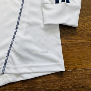 May include: White long-sleeve athletic shirt with navy blue stitching and accents. Features the text "CLIMACOOL" and navy blue stripes on the sleeve cuff. The shirt is made of a lightweight, breathable material.