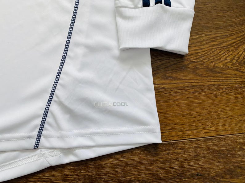 May include: White long-sleeve athletic shirt with navy blue stitching and accents. Features the text "CLIMACOOL" and navy blue stripes on the sleeve cuff. The shirt is made of a lightweight, breathable material.