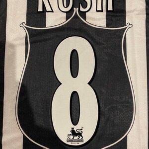 Może przedstawiać: Czarno-biała koszulka piłkarska w paski z napisem "RUSH" u g&oacute;ry. Duża biała cyfra "8" jest wyśrodkowana na czarnej tarczy z logo Premier League. Klasyczny sportowy design.