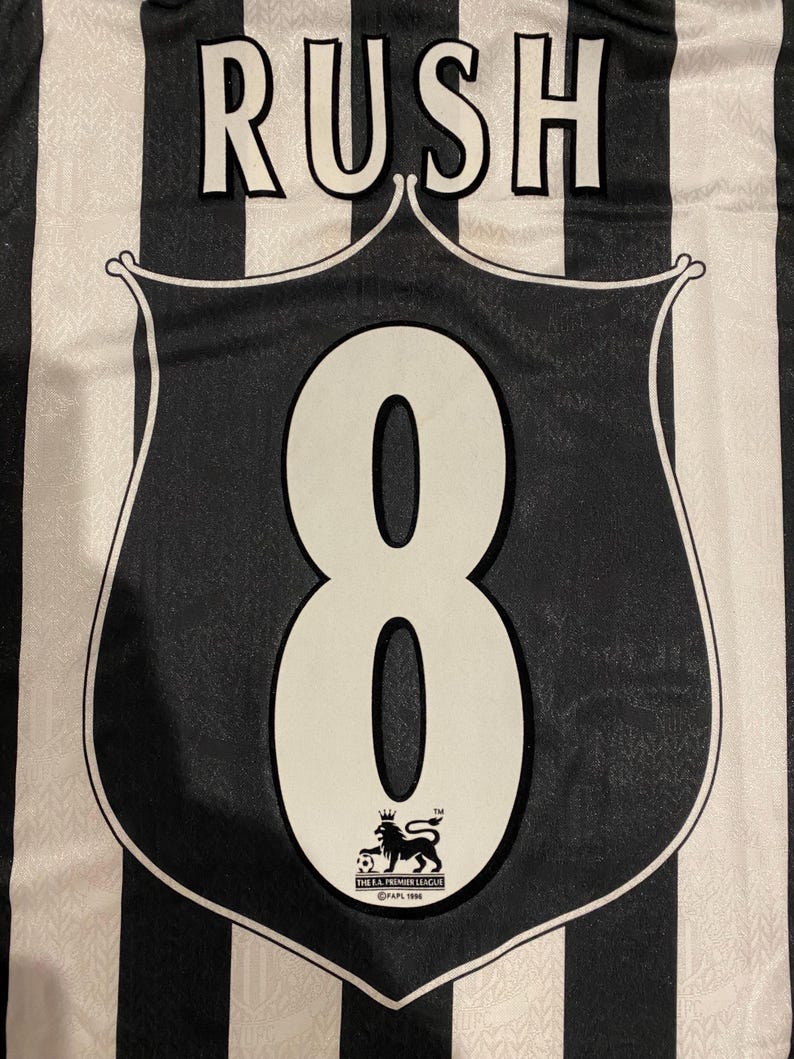 Może przedstawiać: Czarno-biała koszulka piłkarska w paski z napisem "RUSH" u g&oacute;ry. Duża biała cyfra "8" jest wyśrodkowana na czarnej tarczy z logo Premier League. Klasyczny sportowy design.