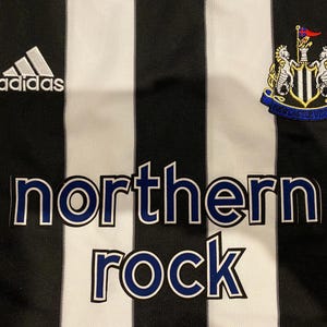 May include: Close-up of a black and white striped jersey. The Adidas logo is visible on the upper left. The words "northern rock" are printed in blue letters. A team crest is on the upper right.