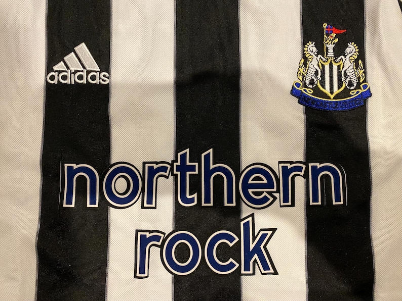 May include: Close-up of a black and white striped jersey. The Adidas logo is visible on the upper left. The words "northern rock" are printed in blue letters. A team crest is on the upper right.