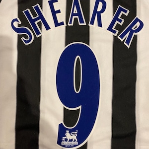 May include: Close-up of a black and white striped soccer jersey. The name "SHEARER" is arched across the top in blue letters with white outlines. Below is a large blue number "9" and a Premier League logo.