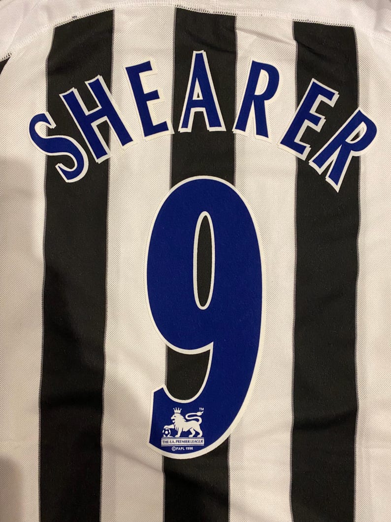 May include: Close-up of a black and white striped soccer jersey. The name "SHEARER" is arched across the top in blue letters with white outlines. Below is a large blue number "9" and a Premier League logo.