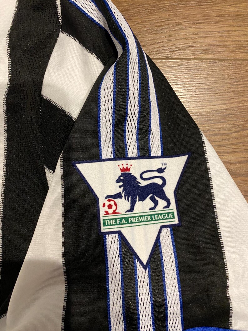 May include: Black and white striped jersey with a Premier League logo. The jersey features a triangular patch with a lion and soccer ball emblem, and the text "THE F.A. PREMIER LEAGUE". The jersey has blue accents.