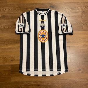 Może przedstawiać: Czarno-biała koszulka piłkarska w paski Newcastle United z logo Newcastle Brown Ale i marką Adidas. Koszulka ma kr&oacute;tkie rękawy i okrągły kołnierz. Widoczne jest logo Premier League.
