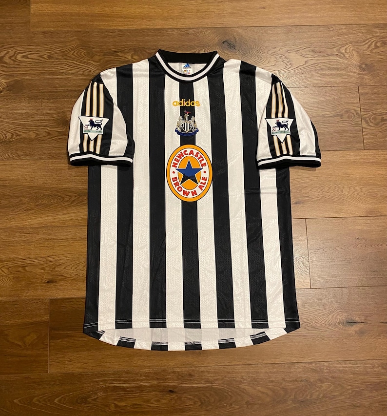 Może przedstawiać: Czarno-biała koszulka piłkarska w paski Newcastle United z logo Newcastle Brown Ale i marką Adidas. Koszulka ma kr&oacute;tkie rękawy i okrągły kołnierz. Widoczne jest logo Premier League.