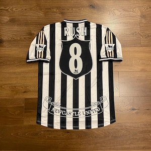 Może przedstawiać: Czarno-biała koszulka piłkarska Newcastle United w paski z nazwiskiem "RUSH" nad numerem "8". Koszulka ma logo drużyny na rękawach i napis "Newcastle United" na dole.