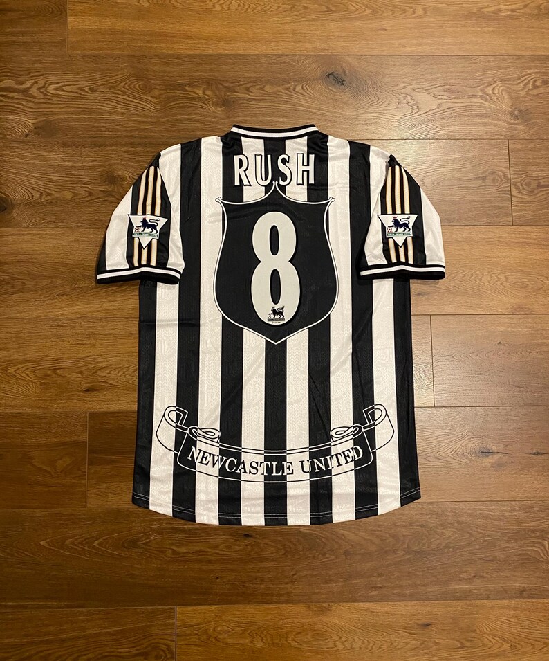 Może przedstawiać: Czarno-biała koszulka piłkarska Newcastle United w paski z nazwiskiem "RUSH" nad numerem "8". Koszulka ma logo drużyny na rękawach i napis "Newcastle United" na dole.