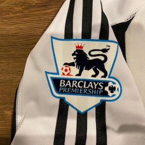 May include: Close-up of a white and black soccer jersey. The jersey features black vertical stripes and a Barclays Premiership logo with a lion and soccer ball. The jersey is made of a breathable fabric.