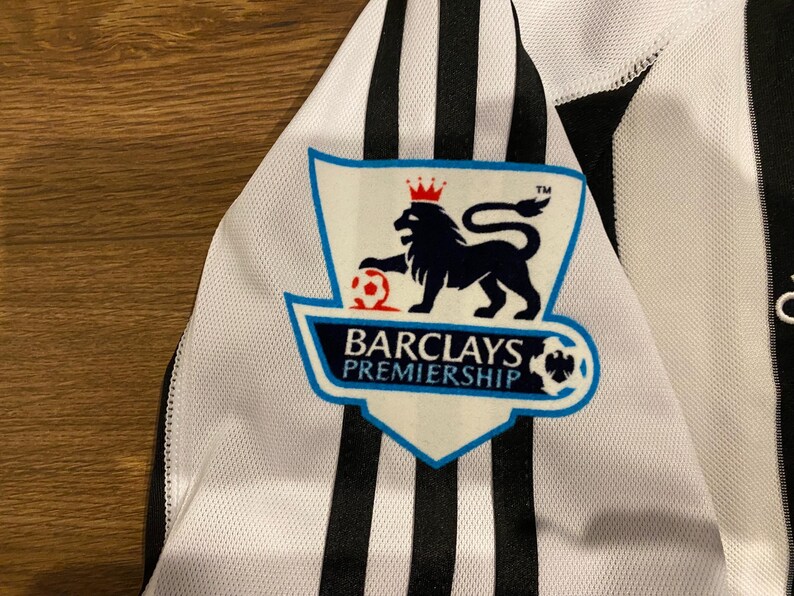 May include: Close-up of a white and black soccer jersey. The jersey features black vertical stripes and a Barclays Premiership logo with a lion and soccer ball. The jersey is made of a breathable fabric.