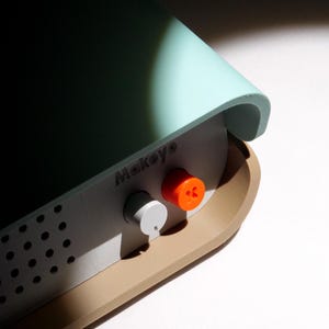May include: A close-up of a MakeyO device. The device has a light blue top and a tan base. It features two knobs, one white and one orange, and the word "Makeyo" is printed on the front.