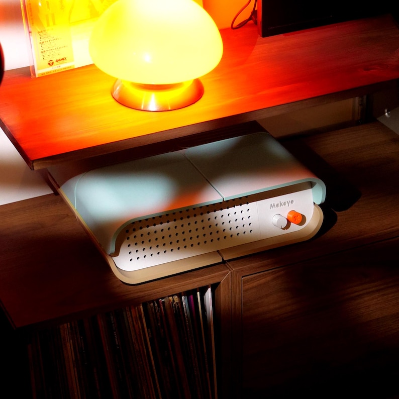 May include: A retro-style audio device with a white and light blue casing, featuring a perforated front panel and an orange knob. The word "Mekeye" is printed on the front. A yellow mushroom-shaped lamp sits above the device on a wooden surface.