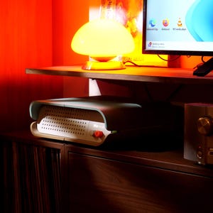 May include: A retro-style entertainment console with a perforated front panel and a red button. The console is teal and white, with a curved design. A yellow mushroom lamp sits on a shelf above the console, next to a computer monitor.