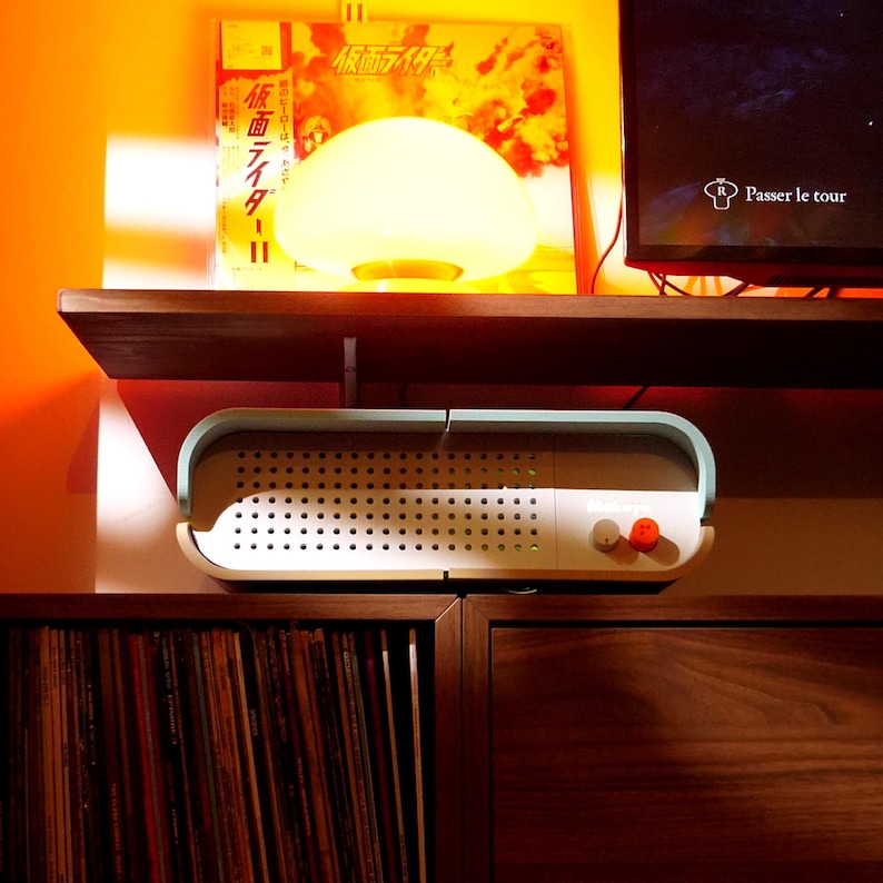 May include: A white and light blue retro-style speaker with a perforated front panel and two control knobs. A glowing mushroom-shaped lamp sits on a shelf above the speaker, with a Japanese album cover in the background.