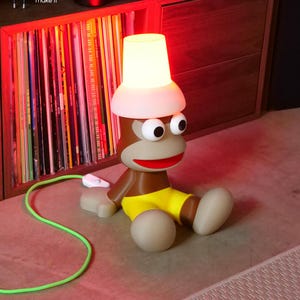 Może przedstawiać: Brązowa lampa w kształcie małpy z dużymi białymi oczami, czerwonymi ustami i żółtymi spodenkami. Lampa posiada świecący biały klosz w kształcie wiadra na głowie oraz jaskrawozielony przewód zasilający z białym przełącznikiem. Tekst: APE Dream it print it make it oraz Makeyo.