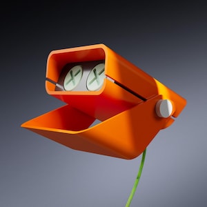 May include: An orange, abstract sculpture with a rectangular top section containing two circular, light green elements with white cross-shaped designs. The sculpture has a lower, angled section and a white circular detail. A green cord extends from the base.