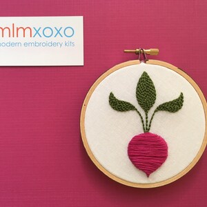 May include: A close-up of a finished embroidery hoop with a pink radish and green leaves stitched on white fabric. The hoop is made of wood and has a diameter of approximately 6 inches.