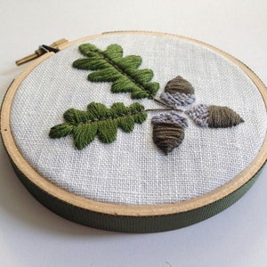 May include: Embroidered hoop art featuring three acorns and two oak leaves in shades of green and gray on a white linen background. The hoop is made of wood and has a green ribbon.