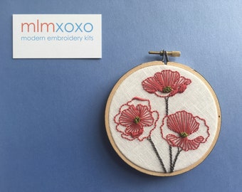 May include: A close-up of a finished embroidery hoop with three red poppies on a white background. The hoop is made of wood and has a black embroidery needle.