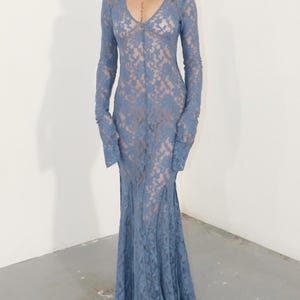 May include: A floor-length, long-sleeved dress crafted from blue floral lace. The garment features a deep V-neckline, a fitted bodice, and a flared skirt that extends to the floor. The lace fabric has a semi-sheer quality, revealing a neutral-toned lining underneath.