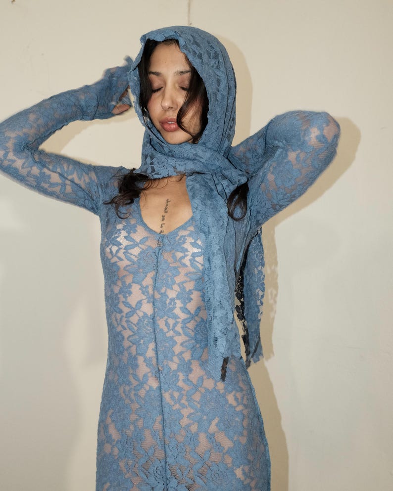 May include: A dusty blue lace dress featuring long sleeves with integrated fingerless gloves and a matching hooded scarf. The garment is crafted from a floral patterned lace fabric that provides a semi-sheer appearance throughout the entire piece.
