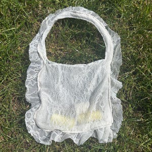 May include: White lace handbag with a rectangular body and a rounded handle. The bag is made of delicate lace with a ruffled edge. The bag is sitting on a bed of green grass.