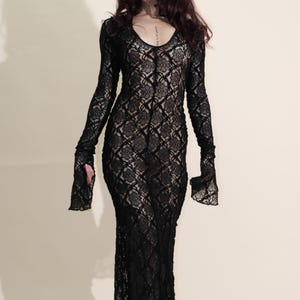 May include: A long, black lace dress with a V-neck and long sleeves. The dress has a fitted silhouette and a flared hem. The sleeves have a bell-like shape at the wrists. The dress is made of a delicate, see-through lace fabric.