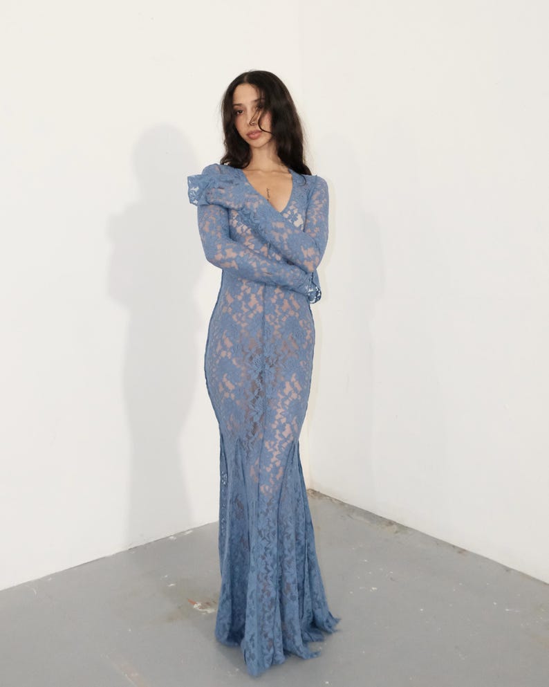 May include: A floor-length, long-sleeved dress crafted from blue lace fabric. The garment features a deep V-neckline and a fitted silhouette that flares slightly at the hem, showcasing a detailed floral lace pattern throughout the entire piece.