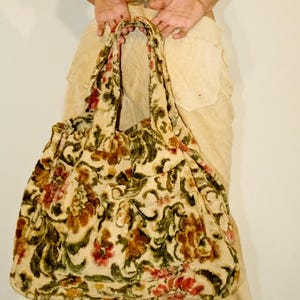 May include: A large, soft tote bag with a floral pattern in shades of cream, brown, red, and green. The bag has a rounded shape and two sturdy handles. The fabric appears to be plush or textured.
