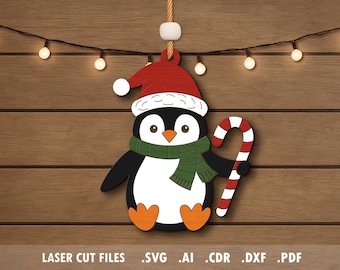 May include: A wooden Christmas ornament featuring a cartoon penguin wearing a Santa hat and green scarf, holding a candy cane. The penguin is black, white, and orange. The background is a wooden wall with string lights. Text at the bottom reads "LASER CUT FILES".