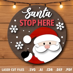 May include: A circular wooden sign with a Santa Claus design. The sign displays the words "Santa STOP HERE" in white and red, with snowflake accents. The Santa illustration has a red hat and suit, white beard, and black gloves. The sign is set against a wooden backdrop.