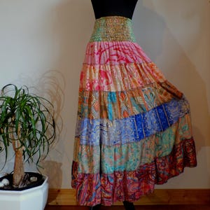 Pu&ograve; includere: Una gonna lunga a pi&ugrave; strati con un colorato design patchwork. La gonna presenta un'ampia fascia elastica in vita e una silhouette fluida. Il tessuto mostra una variet&agrave; di motivi e colori, tra cui rosa, blu, arancione e verde. La gonna &egrave; esposta su un manichino nero.