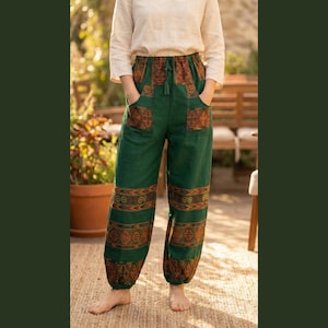 Op de afbeelding: Groene broek met bruine geometrische patronen op de zakken en de onderbenen. De broek heeft een taille met trekkoord en zakken. De persoon op de foto draagt een crèmekleurig shirt met lange mouwen en staat buiten.